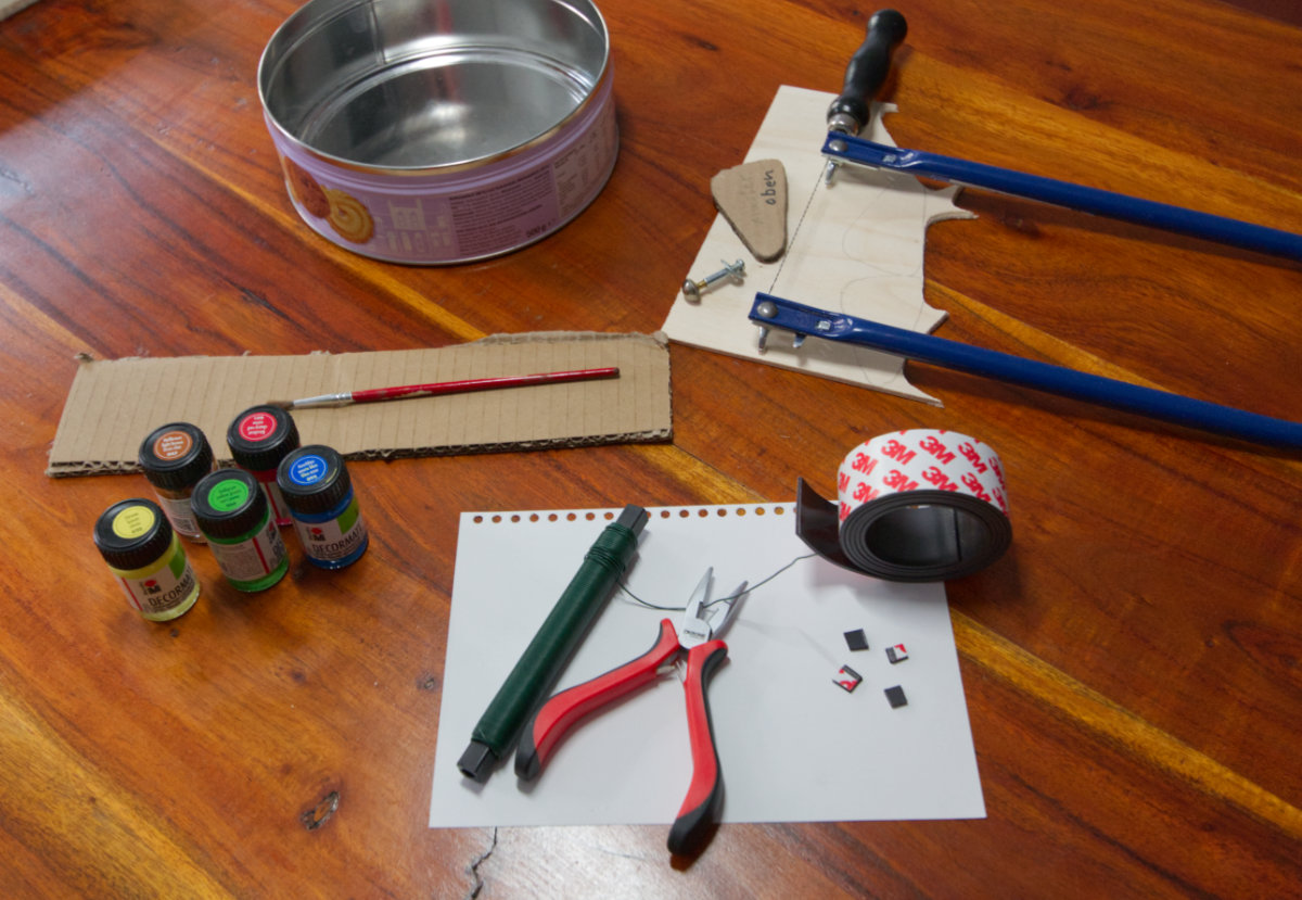 Handicraft accessories on a wooden table top: pliers, brushes, 5 paint flasks, binding wire, cardboard, plywood, a fretsaw, a long screw with nut